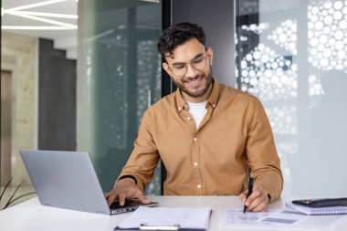 Dizüstü bilgisayarda çalışan ve modern bir ofiste not alan gülümseyen bir iş adamı. İş yerindeki görevleri yönetirken profesyonel, üretken ve odaklı.