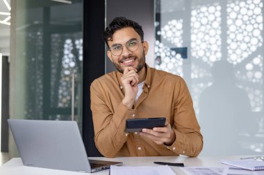 Modern ofis ortamında hesap makinesi kullanarak masa başında çalışan gülümseyen bir adam. Dizüstü bilgisayar ve evraklar masada görünür..