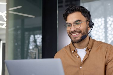 Gülümseyen müşteri hizmetleri temsilcisi kulaklık ve gözlük takıyor, modern bir ofis alanında dizüstü bilgisayarla çalışıyor..