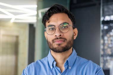 Modern ofis ortamında duran mavi gömlekli, gözlüklü ve sakallı kendinden emin genç bir adam..