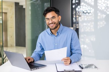 Mavi gömlekli gülümseyen işadamı laptopta çalışıyor ve elinde tablet ve kağıtlarla bir ofis masasında belgeleri inceliyor..
