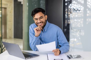 Sırıtkan işadamı masamda dizüstü bilgisayar ve tabletle belge okuyor. Modern işyeri ortamını ve verimli iş faaliyetlerini gösteren profesyonel ofis ortamları.