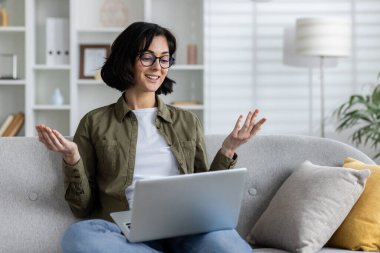 Günlük giysiler içinde gülümseyen genç bir kadın evdeki kanepede oturuyor, kucağında dizüstü bilgisayarını tutuyor ve elleriyle işaret ederken video görüşmesinde konuşuyor..