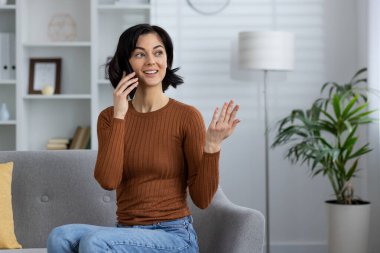 Modern bir oturma odasında otururken telefonuyla neşeli bir konuşma yapan bir kadın. Mutlu ve nişanlı görünüyor..