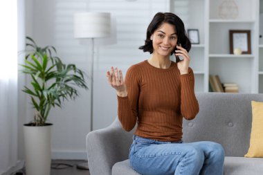 Oturma odasındaki kanepede otururken telefonda konuşan gülümseyen kadın. Sıradan ve dostane bir atmosfer yakalar..