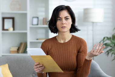 Elinde bir zarf tutarken kafası karışmış bir kadın, rahat bir evde oturma odasında oturuyor..