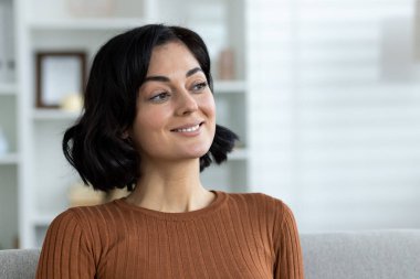 Oturma odasında kanepede oturan gülümseyen kadın düşünceli ve mutlu görünüyor. Konforlu ev ortamı