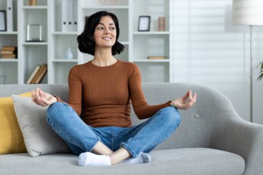 Oturma odasında kanepede oturmuş meditasyon yapan genç bir kadın. Rahat ve huzurlu bir ortam..