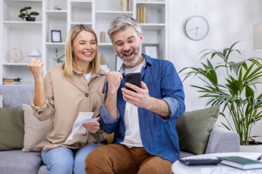 Joyful couple celebrating good news with a smartphone and document while sitting on a couch at home. clipart