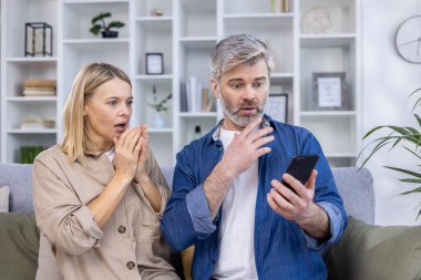 Orta yaşlı bir çift, modern bir oturma odasında kanepede otururken akıllı bir telefona bakıyorlar..