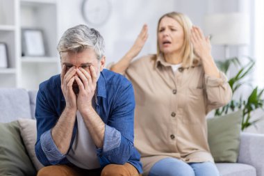 Ortağı oturma odalarında ateşli bir tartışma sırasında arka planda hareketler yaparken sinirli adam ellerini yüzünü kapatıyor..