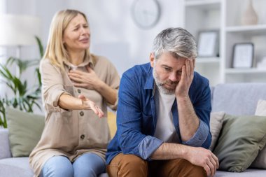 Orta yaşlı çift, duygusal stres ve gerilim göstergesi olarak evde çatışmayla karşı karşıya. Kadın duygusal olarak hareket ederken, erkek üzgün ve endişeli görünüyor..