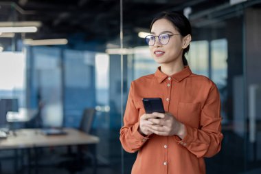 Modern ofiste akıllı telefon kullanan Asyalı bir iş kadını. Profesyonel güveni ve iş ortamını yakalar. İş, teknoloji ve iletişim kavramları için ideal.