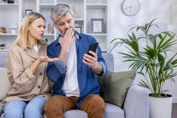 Беспокойная пара, сидящая на диване, реагируя на неожиданные новости со смартфона в своей гостиной.