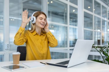 Woman wearing headphones, listening to music, and enjoying a video call while sitting at her office desk with a laptop.