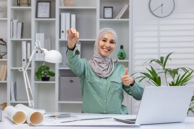 Confident female realtor wearing a hijab holds house keys and gives a thumbs up while working in a modern office. Concept of success, real estate, and professional achievement.