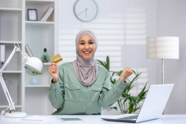 Smiling woman wearing hijab holding a credit card while working on a laptop at a modern home office. Successful online shopping and digital transactions concept.