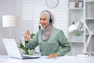 Smiling woman wearing a hijab using a laptop for a video call while working from home in a modern office setup.