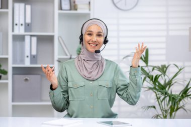 Smiling customer support representative wearing hijab and headset, working in a modern office setting with bookshelves and plants.