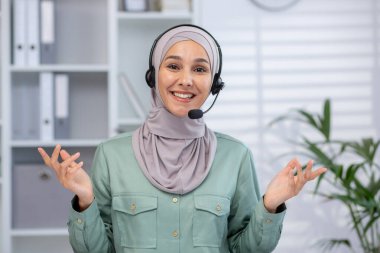 Smiling woman wearing hijab with headset giving online presentation in office. Professional support worker interacting during virtual meeting in a modern workplace environment.