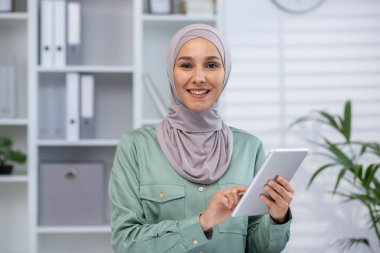 Smiling woman in hijab holding and using a digital tablet in a modern office environment with shelves and plants in the background.