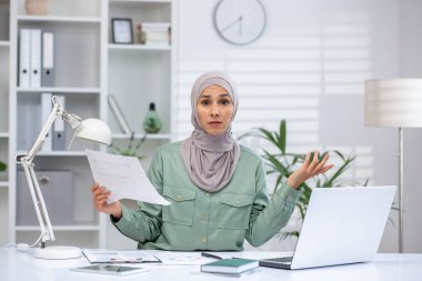 Frustrated businesswoman wearing hijab holding documents, attending an online meeting in a modern office. Concept of work stress, confusion, and business challenges.