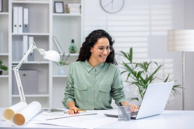 Hispanic woman architect working on a laptop in a modern office, surrounded by plants and office supplies, looking happy and focused.