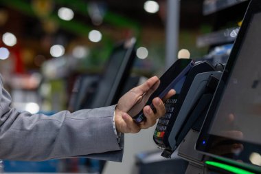 Image shows a person using a smartphone for NFC Near Field Communication mobile payment on a point-of-sale terminal in a retail environment. The focus is on the technology and convenience.