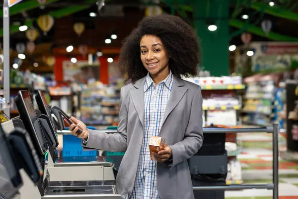 stock image In a grocery store, a young woman using a selfcheckout machine as she holds a smartphone and a grocery item. This scene depicts a modern shopping experience with digital technology