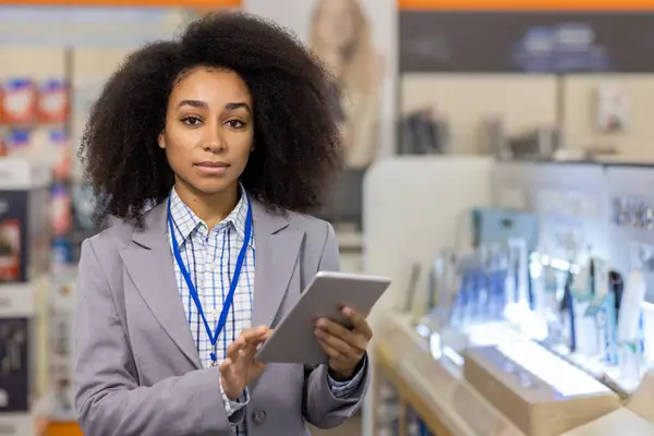 In an electronics store, a confident professional woman with a badge holds a tablet, surrounded by shelves of gadgets. She symbolizes expertise and modern business in retail technology