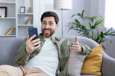Man enjoying a video call on his smartphone, sitting comfortably on a couch at home with a cheerful expression.