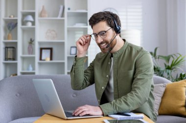 Confident man working from home using a laptop and headset, smiling pleasantly. Home office setup with a cozy background. Concept of remote work and online communication.