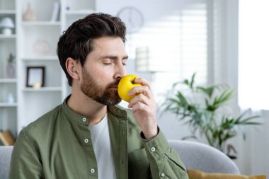 A man with a beard is peacefully smelling a fresh apple while sitting in a cozy living room.