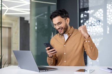 Modern bir ofiste başarıyı kutlayan heyecanlı bir adam akıllı telefonuna bakarak ve sevinçle yumruğunu kaldırarak..