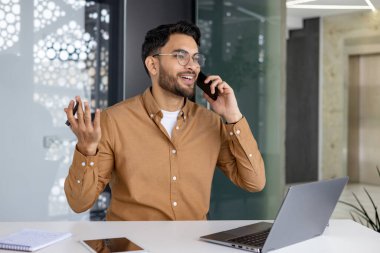 Gözlüklü gülümseyen genç işadamı, modern ofis ortamında dizüstü bilgisayarı üzerinde çalışırken hoş bir telefon görüşmesi yapıyor..