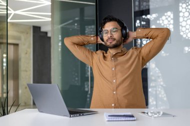 Kulaklık takan genç bir profesyonel, elleri ofiste dizüstü bilgisayar ve defterle dinleniyor. Modern ofis ortamı ve rahat çalışma ortamı.