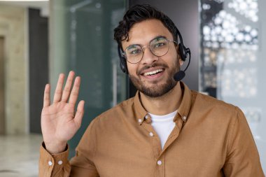 Gözlüklü ve kulaklıklı gülümseyen müşteri hizmetleri temsilcisi modern ofis ortamında kameraya el sallıyor ve bakıyor..
