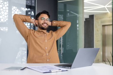 Gülümseyen adam masasında ellerini başının arkasına koyup dinleniyor, modern ofis ortamında işe ara veriyor.