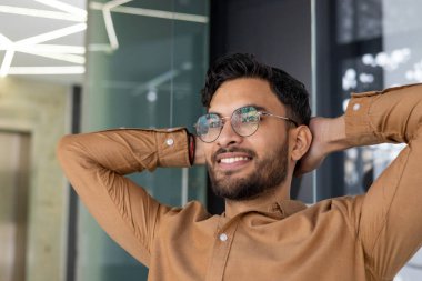 Modern ofis ortamında rahatça gülümseyen gözlüklü genç adam. Kahverengi gömlek, rahat poz ve çağdaş arka plan..