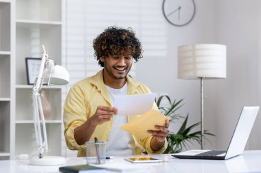 Gülümseyen adam modern bir ofiste masa başında otururken bir mektup açıyor. Olumlu duygular, evrak işleri ve profesyonel çalışma ortamı gösterir.