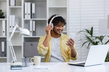 Evde çalışan genç bir adam, kulaklıkla müzik dinlerken akıllı telefonunu kullanıyor, neşeli bir tavır sergiliyor.