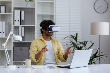 VR kulaklık takan ve ofis masasında sanal çevreyle etkileşime giren bir adam, önünde bir dizüstü bilgisayarla..