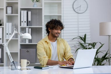 Modern bir ofiste dizüstü bilgisayarda çalışan gülümseyen bir adam. Çalışma alanı kitap rafları, bir lamba ve masa gereçleriyle düzenlenir..