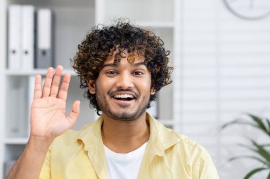 Görüntülü arama sırasında gülümseyen genç adam el sallıyor. Ev ofis ortamında arkadaşça ve mutlu bir etkileşim..