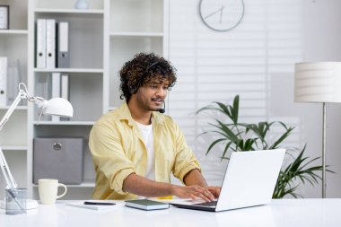 Kulaklıklı genç adam ofis masasında dizüstü bilgisayarda çalışıyor, modern ve parlak bir ofis alanında verimli ve odaklanmış bir çalışma ortamı yaratıyor..