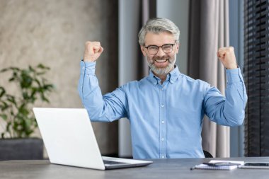Modern bir ofiste masa başında oturan başarılı bir işadamı bir dizüstü bilgisayar kullanarak başarısını neşeyle kutlar.
