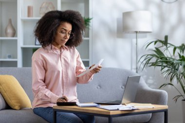 Kıvırcık saçlı ve gözlüklü bir kadın gri bir kanepeye oturmuş hesap makinesi kullanıyor, elinde kağıt, dizüstü bilgisayarı ve masada notları var. Odaklanmış görünüyor, ev ofisinden çalışıyor.