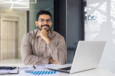 Bir iş adamı masa başında oturuyor ve modern bir ofis alanında dizüstü bilgisayar üzerinde çalışıyor. Masa hesap tabloları ve çizelgeler de dahil olmak üzere belgelerle dolu. Kişi kendinden emin bir şekilde gülümsüyor..