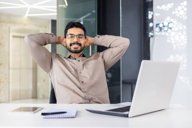 Bir adam modern ofisinde dizüstü bilgisayarı, not defteri ve telefonuyla oturuyor. Rahat ve rahat görünüyor, profesyonel bir ortamda bir anlık rahatlığın tadını çıkarıyor..