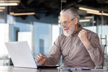 Keyifli bir kıdemli işadamı çağdaş bir ofiste başarısını bir dizüstü bilgisayarın önünde kutluyor.
